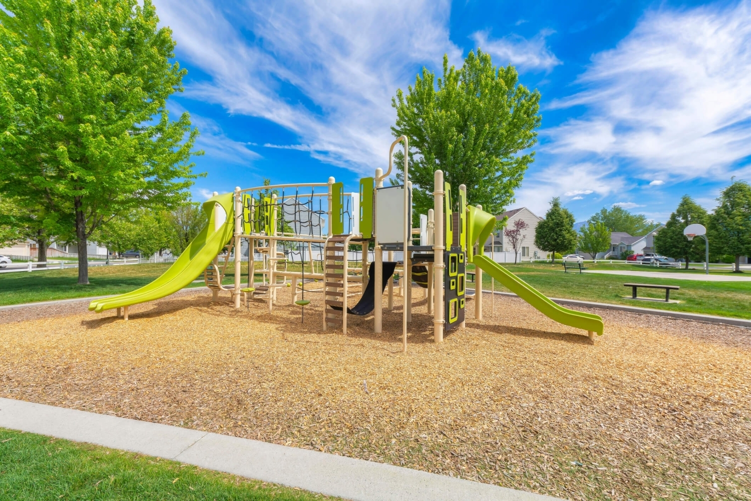 A green anbrown-coloreded playground in a public play area surrounded by trees and a blue sky.