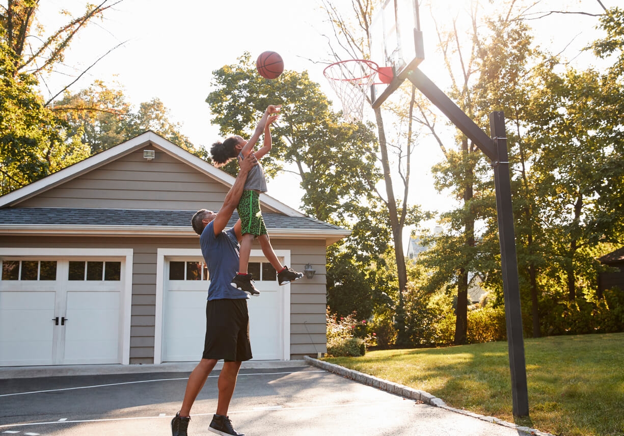 A father holding his son up as he scores a basketball goal in their driveway.