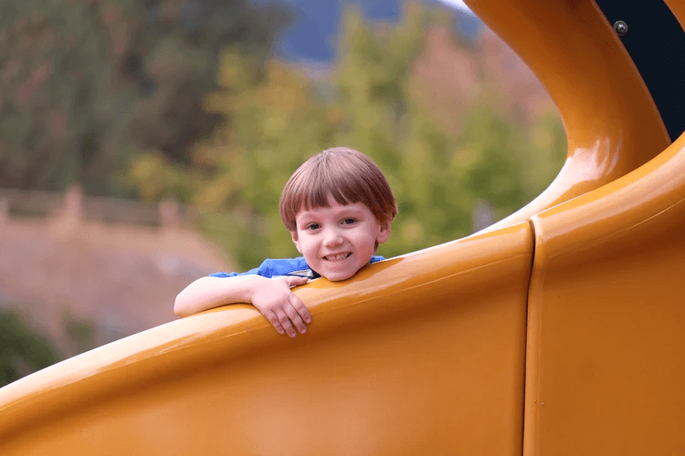 A happy child smiles while sitting on the end of a slide that is part of play equipment.