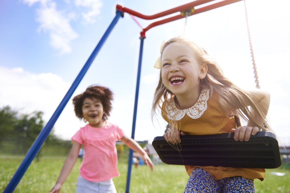 Two little girls laughing and playing on a red and blue swing set.